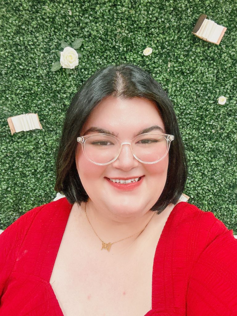 A woman with short dark brown hair and glasses smiles at the camera in front of a greenery background. She is wearing a red shirt.