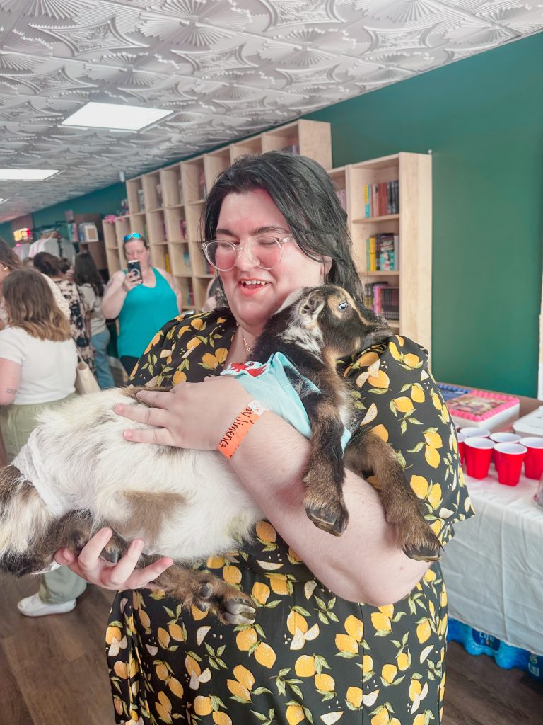 A woman (Bree) with short dark hair and a blouse with lemons on it laughs in joy as she holds a baby goat. Behind her are various other people and bookshelves.