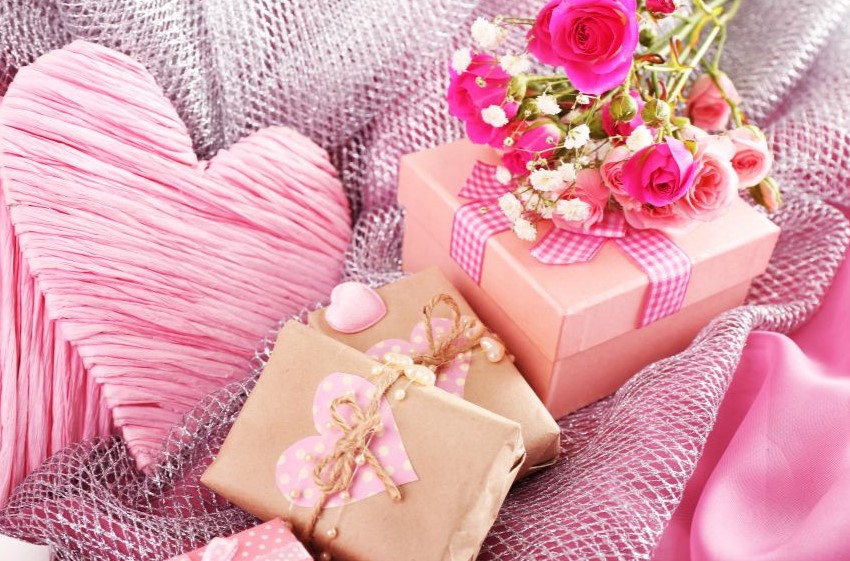 A pink heart, pink box with pink roses on top, and brown packages with pink hearts lying on pink mesh material.