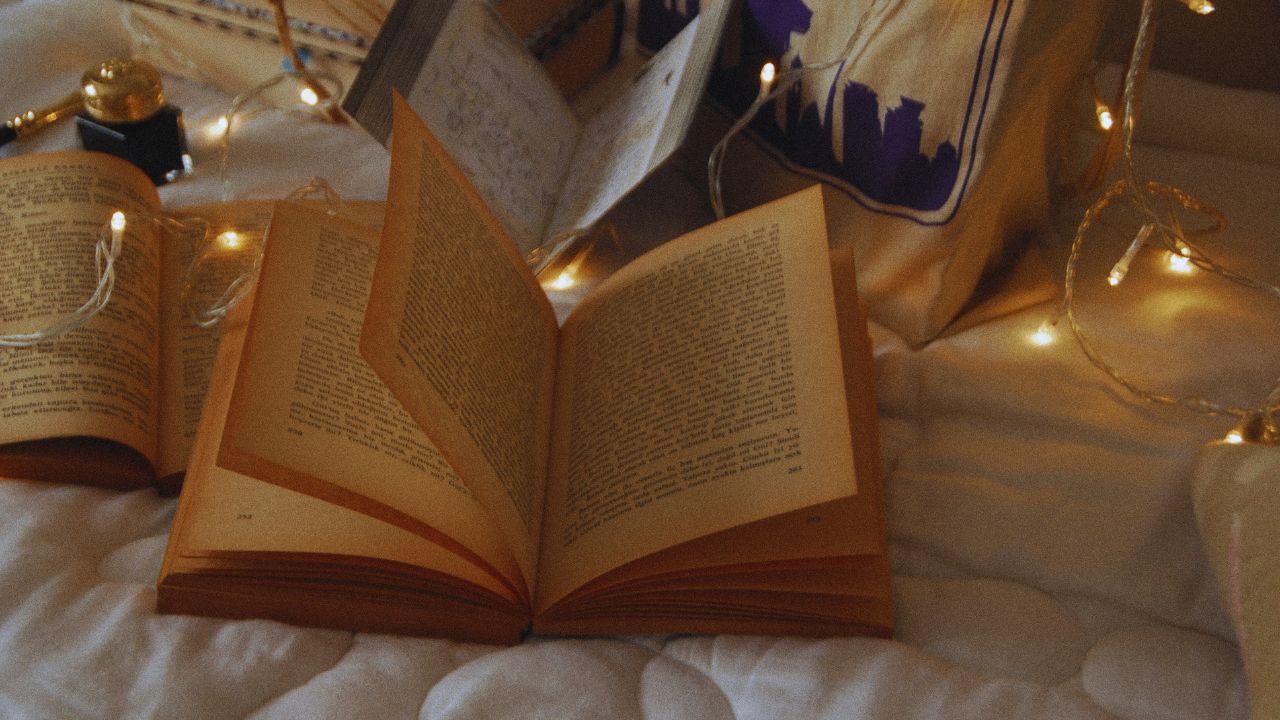 An old book with yellowing pages lies open on a white comforter with a string of warm lights running across the background.