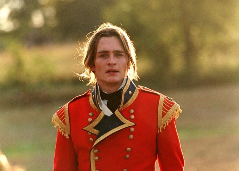 A man with blonde hair pulled back looks very slightly right of the camera from a distance. He is wearing a red British regiment outfit and standing in a green field.