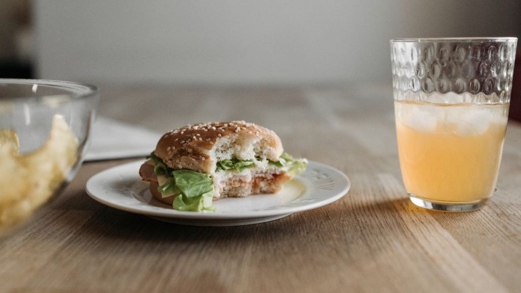 A half eaten sandwich sitting on a plate on a light wooden table. There is a bow of chips half obscured on the left side and a half full glass of orange liquid on the right.