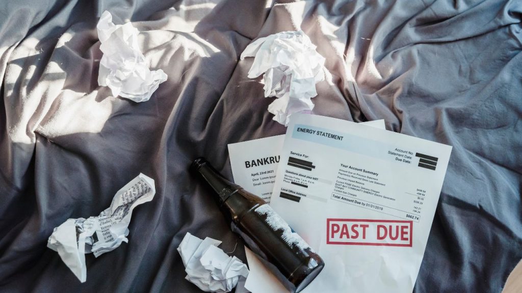 Grey bedsheets with crumpled up papers, bills that say PAST DUE, and a brown bottle with the label ripped off.