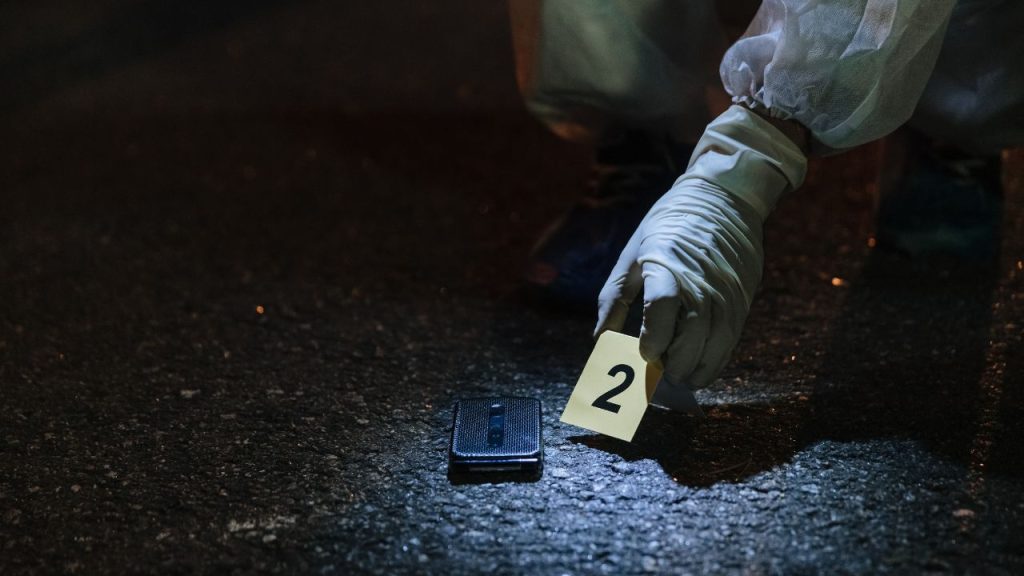 Someone in a white glove and puffy thin white sleeves placing a yellow card that says "2" on it near a phone on the pavement. It looks like a crime scene.
