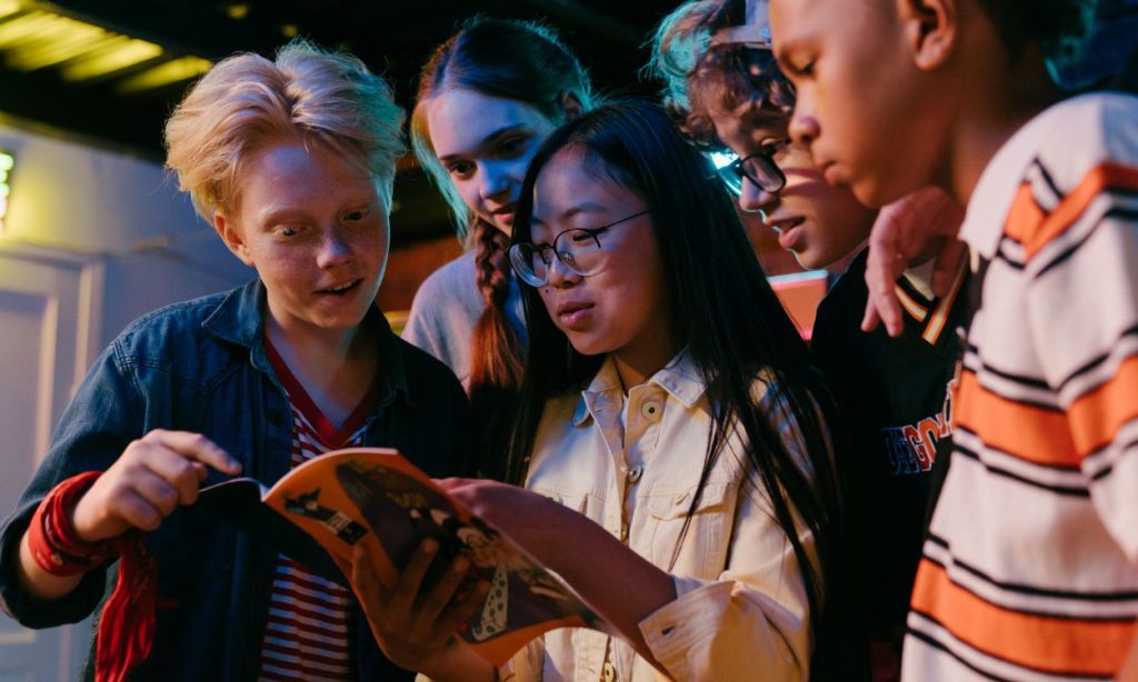 Five friends are huddled around reading an orange book. Two are girls and three are boys.