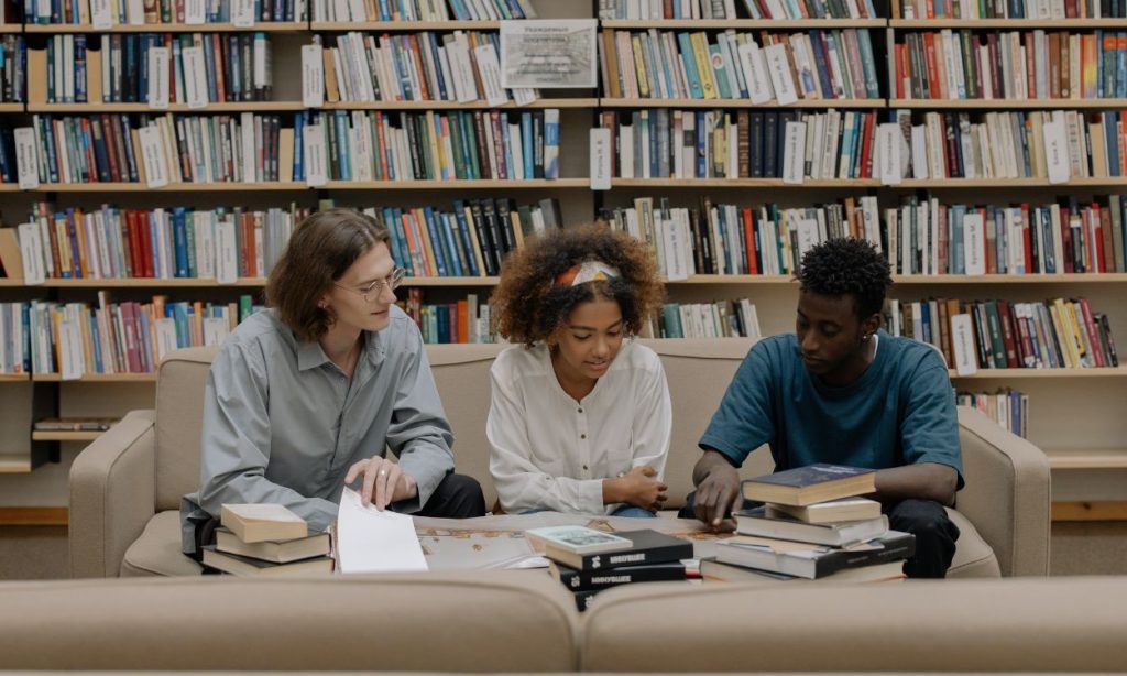 Three young people sitting on a tan couch looking at books on a table. They are in front of a giant bookshelf, presumably at the library.