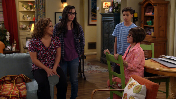 One Day at a Time still with four family members sitting in the kitchen and living room area of their apartment talking to each other.