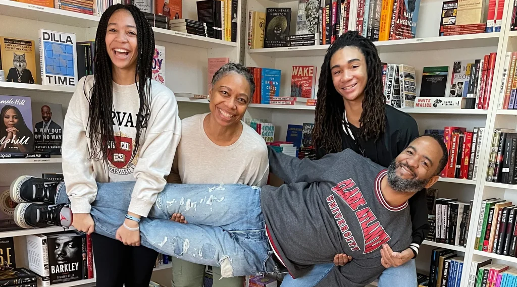 Image depicting the founders of black pearl books, two women and one guy holding a men in a horizontal position, in front of a white bookshelf.