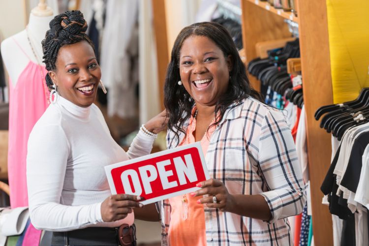 Two smiling women holding an 'Open' sign inside a clothing store, representing successful Black-owned businesses