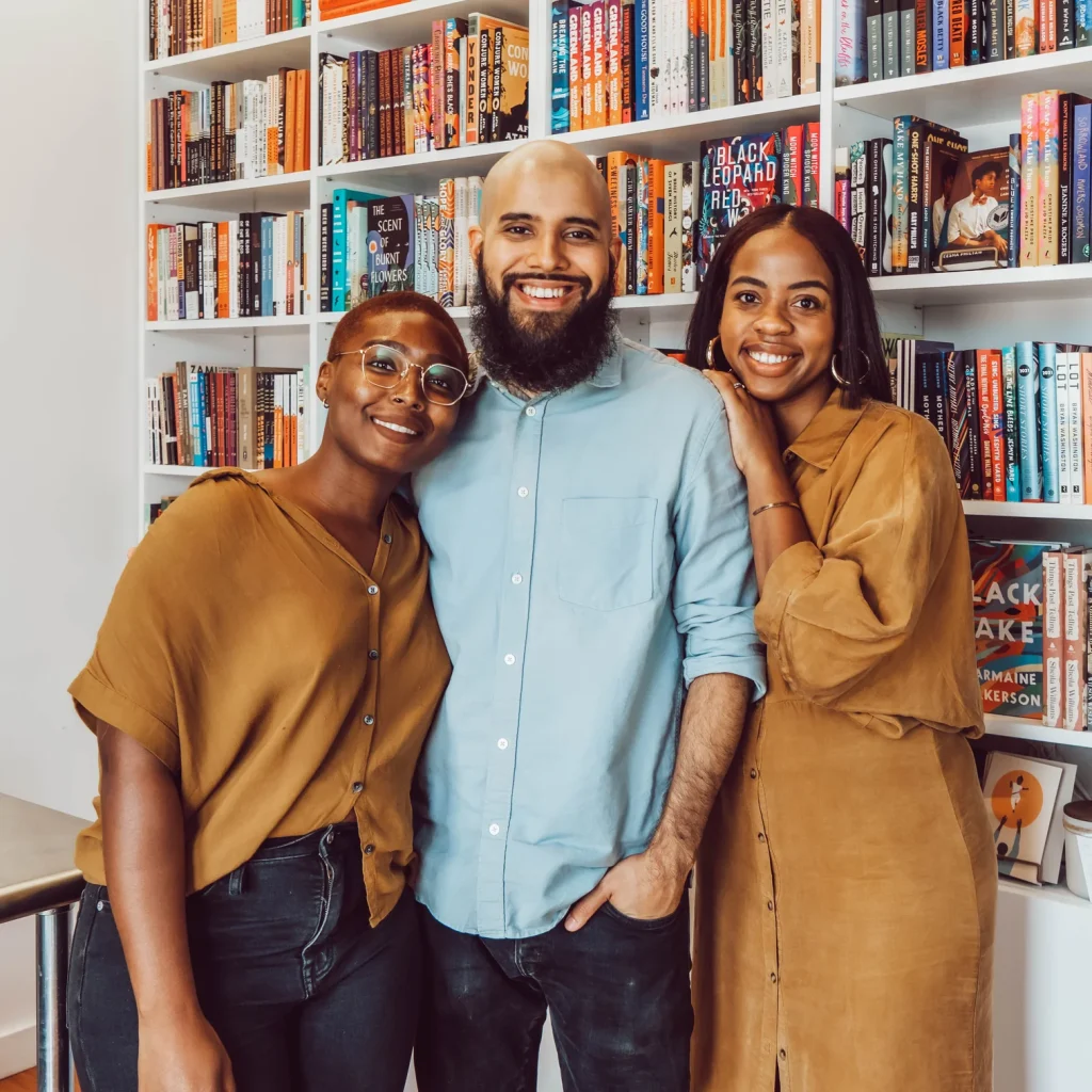 Two women and one men posing for a photo infront of a white shelf filled with books