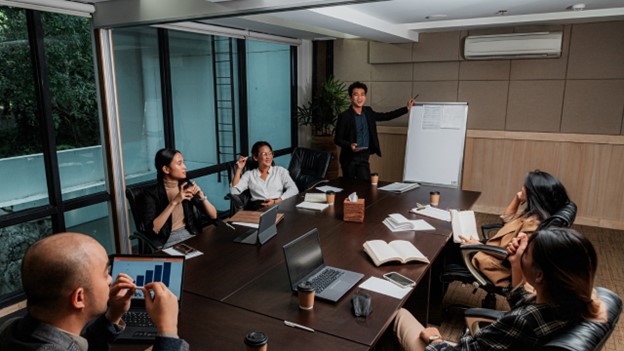 A team of six people in a board meeting. One gives a presentation on a whiteboard, while the other five look up at him.