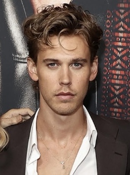 Actor Austin Butler with curly hair and wearing a brown suit with an open white shirt.
