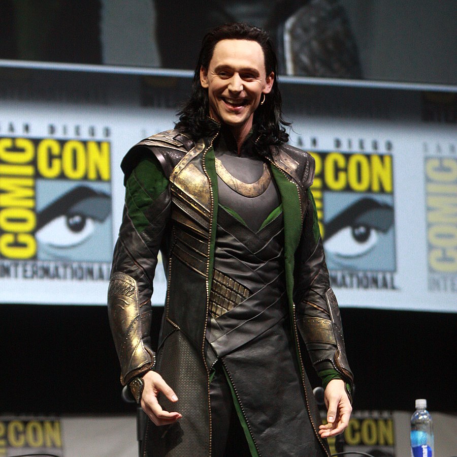 Actor Tom Hiddleston as Loki, God of Mischief, donning his green and gold attire while at Comic Con. He smiling, and his hair is slicked back and down to his shoulders. The "Comic Con" images are set in the background.
