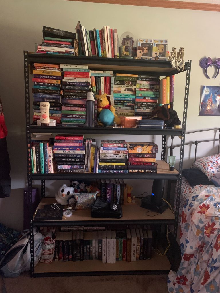A bookcase stacked with books and miscellaneous items, next to a bed with floral sheets.