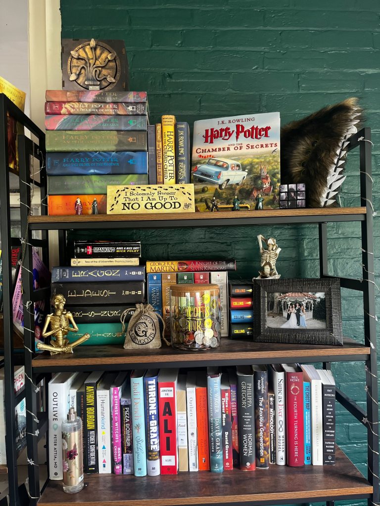 A bookcase full of books and Harry Potter merch against a dark green brick wall.