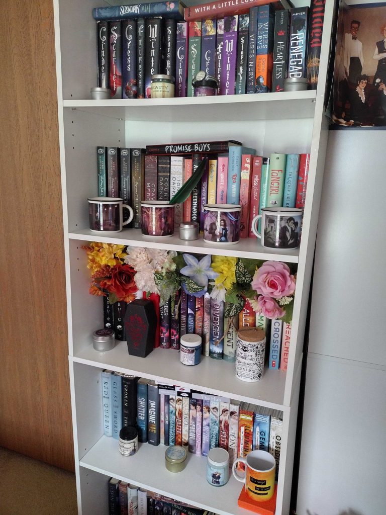 White bookcase full of colorful books, lined up mugs and candles, and flowers.