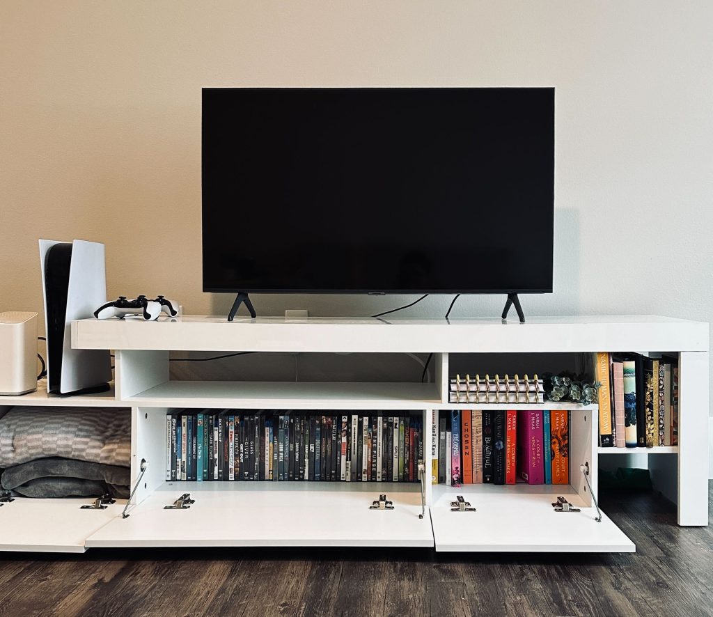 White TV stand stocked with books, DVDs, notebooks, and blankets. Propped on top is a black TV screen, and a PS5 game console and controllers.