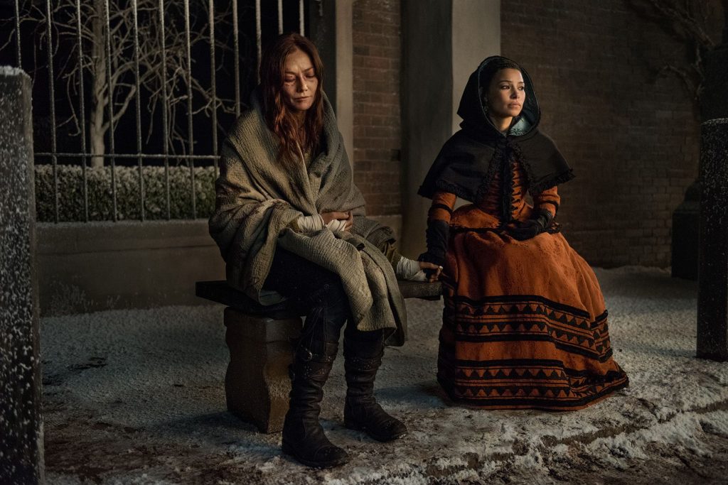 Anne and Max sitting outside. The ground is dusted with snow, and Anne is wrapped in a tan blanket, while Max is bundled in a long-sleeve orange dress and brown hood.