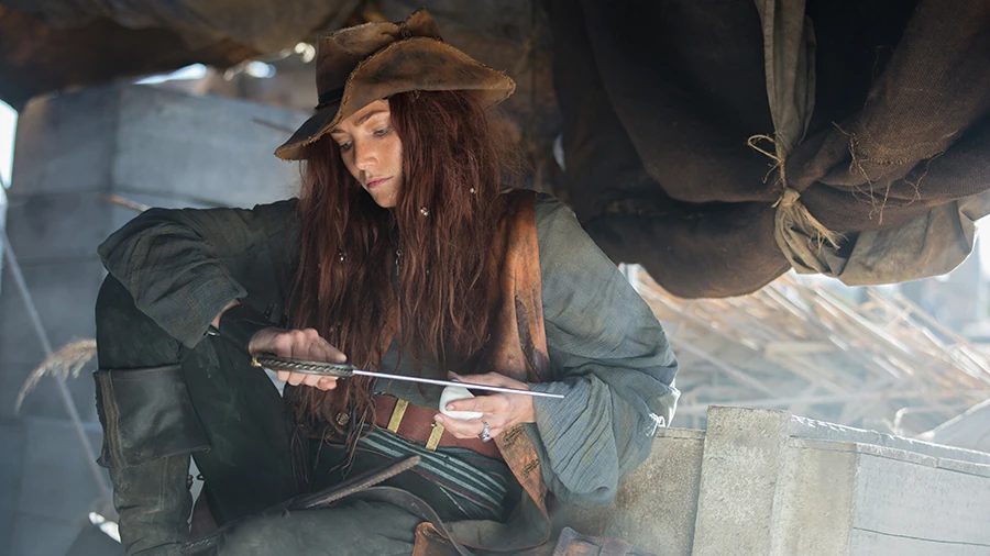 Anne sitting and sharpening a knife with a stone amongst ship cargo. Her long, wavy red hair hangs loose from a worn brown hat.