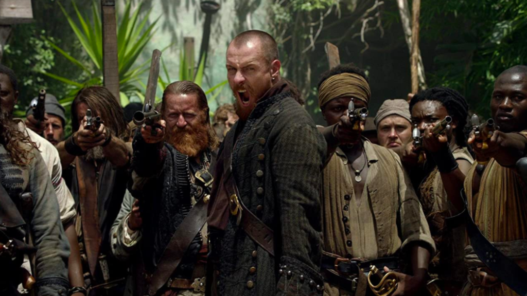 Flint screaming while his crew stands behind him wielding rifles and aiming them straight ahead.