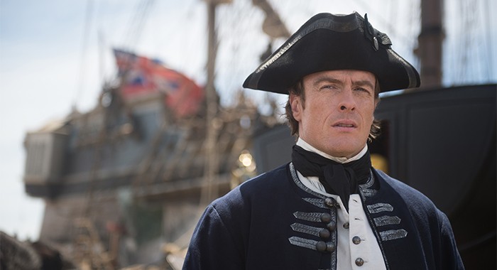 Captain Flint wearing a blue British Royal Navy uniform and standing in front of a Navy ship with a Union Jack flag on it.