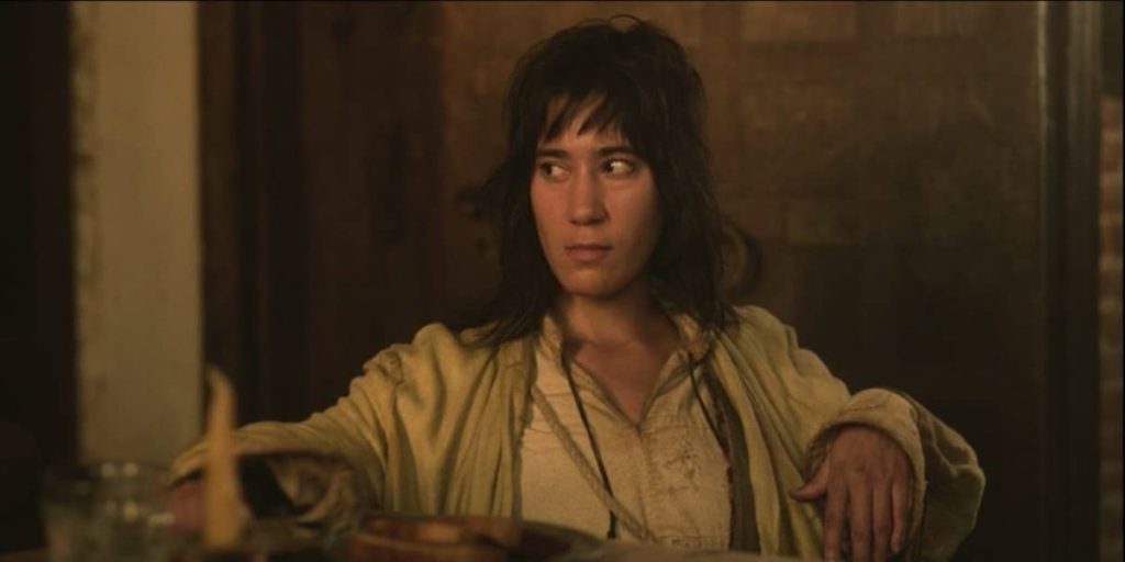 Jim sitting and looking off to the left. Their hair worn down to their shoulders, and they are wearing dirty white tunic.