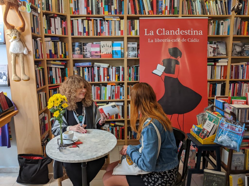 Two women are sitting at a small table in La Clandestina, surrounded by shelves filled with books, with one woman showing a book to the other.