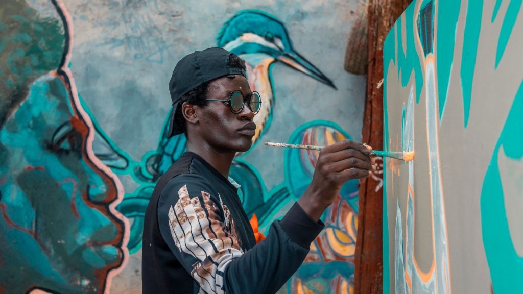 A black person in glasses wearing a backwards cap paints a mural.