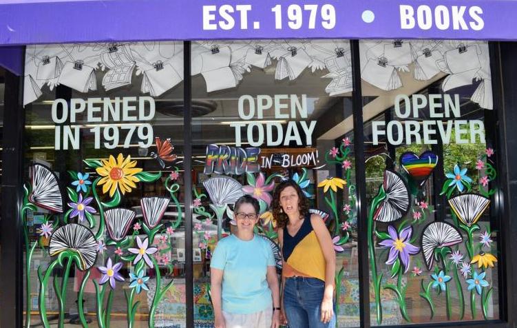 Women & Children First owners Lynn Mooney and Sarah Hollenbeck stand in front of the bookstore windows, which have painted white books hanging from the top and colorful flowers near the bottom with the words "PRIDE IN-BLOOM." The windows read "OPENED IN 1979 | OPEN TODAY | OPEN FOREVER," and the top purple overhead reads "EST. 1979 | BOOKS."