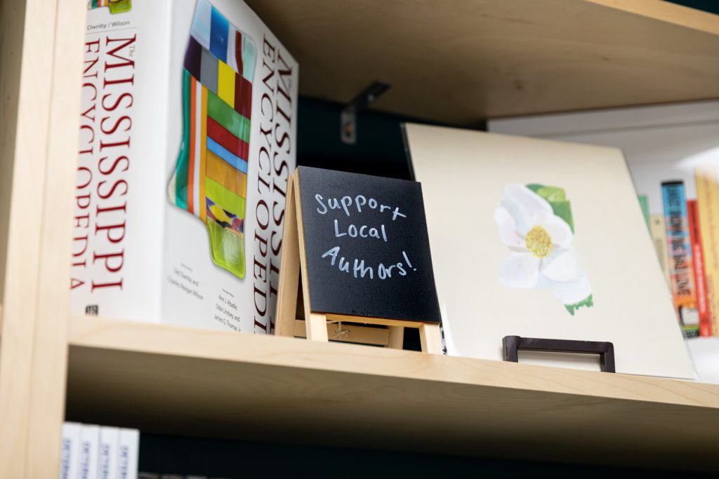 A bookshelf shelf is decorated with a piece of artwork with a white flower, an illustration of books in the background, and a "Mississippi Encyclopedia," along with a small black easel that reads "Support Local Authors!"