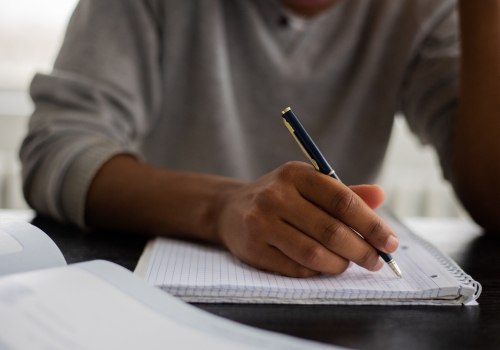 A close-up of a man in a grey jumper, writing on a piece of lined paper with a pen.