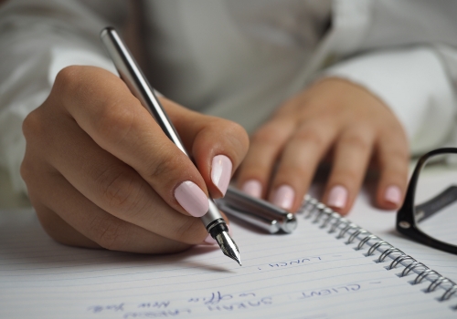 A writer holds a fountain pen poised above a lined notebook. The edge of a pair of glasses and the pen lid lie on the page.