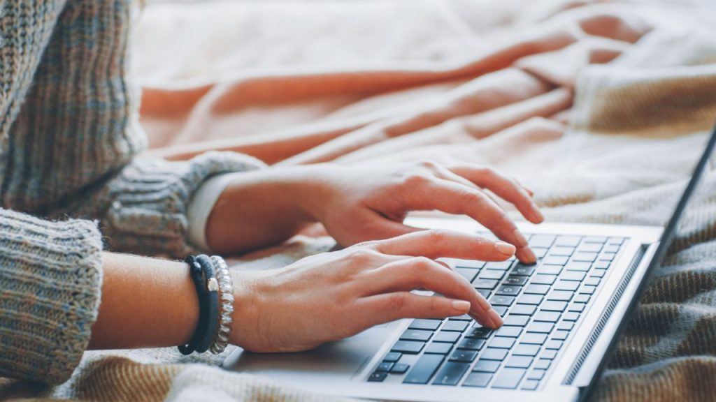 Someone's upper body resting on their elbows as they lean in and type on their laptop. They are sitting on their bed.