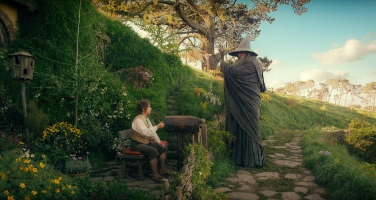 A country side of greenery and flowers. Bilbo is sitting on a bench, smoking a pipe. Gandalf stands over him, with his walking stick and pointy hat, as they converse.