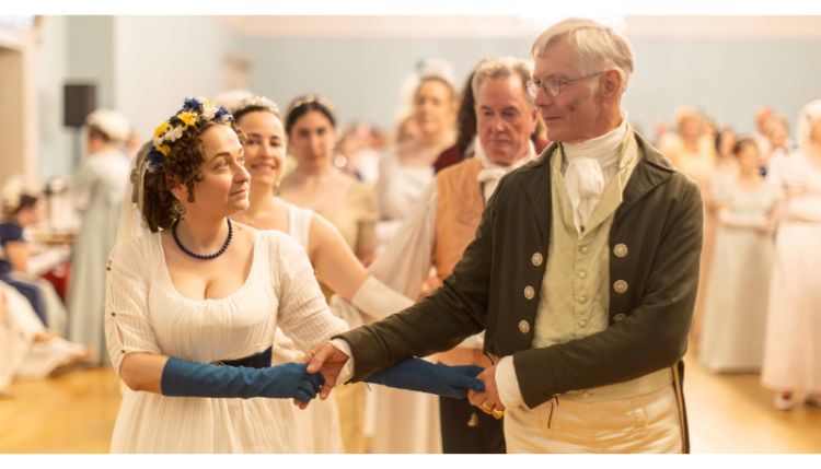 A man and woman dance together dressed up in regency clothes. In the background, others doing the same can be seen