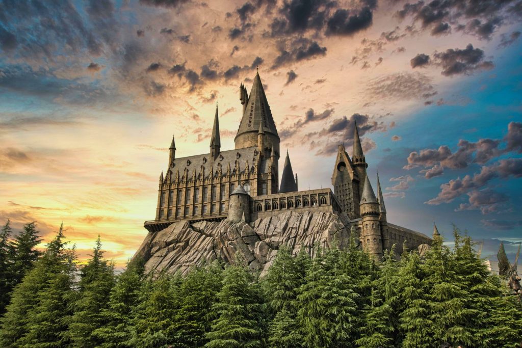 Hogwarts castle built onto a cliff side. In front of it are green trees, and behind it is a blue/yellow/purple sunset.