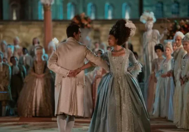 Two people dancing, dressed in regency attire. Other people stand on the periphery, but the focus is on the main couple who are staring at each other.