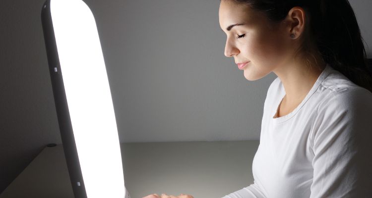 A woman in a white shirt with her eyes closed is basking in the light of a rectangular lamp. The background is gray.