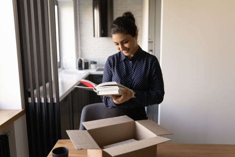 A woman is smiling as she looks at an open book she has taken from a stack of books on a wooden table in her apartment.
