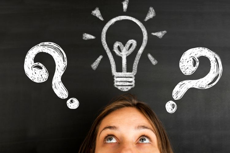 A women´s face in front of a dark board with chalk question marks and a light bulb drawn over her head to show her pondering.