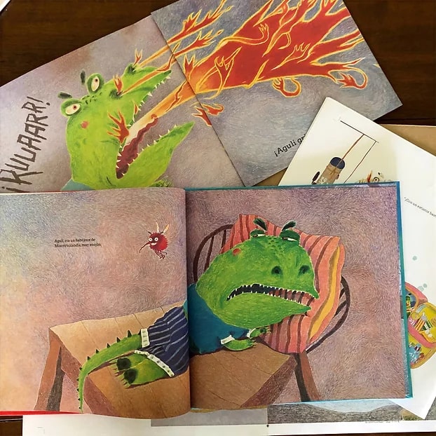 Three open illustrated books from La Cifra Editorial are laid out on a wooden table, each depicting a green lizard spitting fire and waking up in an annoyed mood. The vibrant illustrations capture the dynamic storytelling and artistic style characteristic of La Cifra Editorial's publications