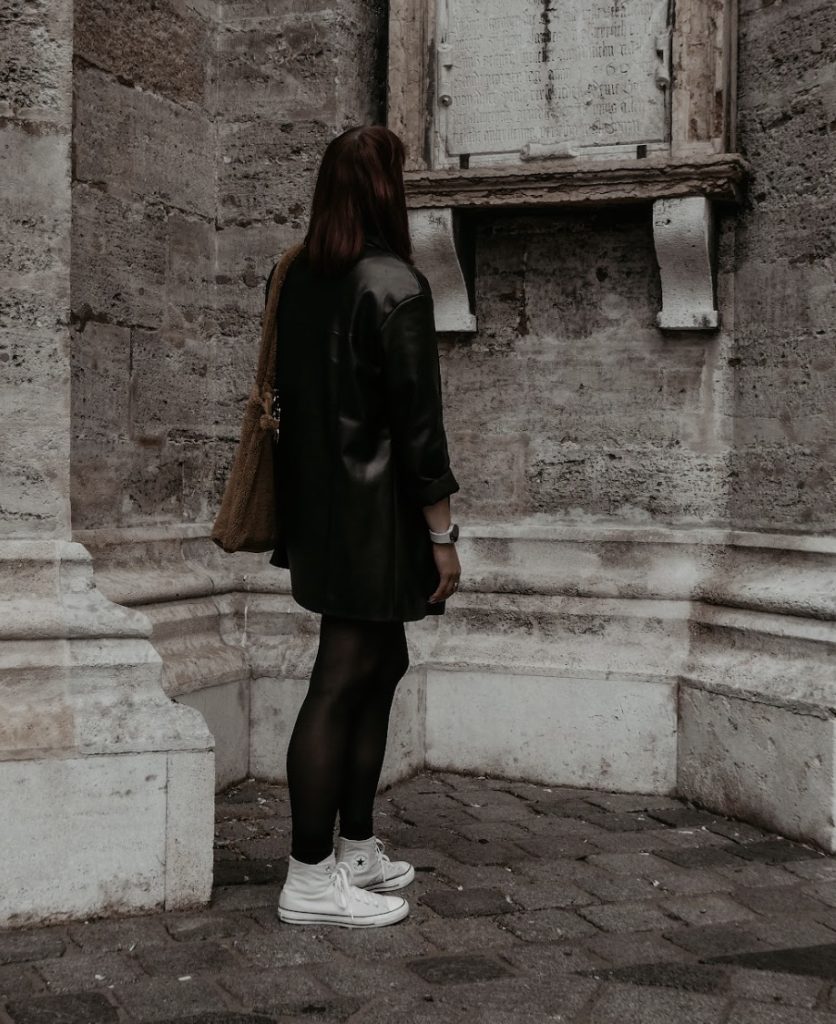Vicky with her back turned to the camera reading an engraving off of an old stone building in a black leather jacket.