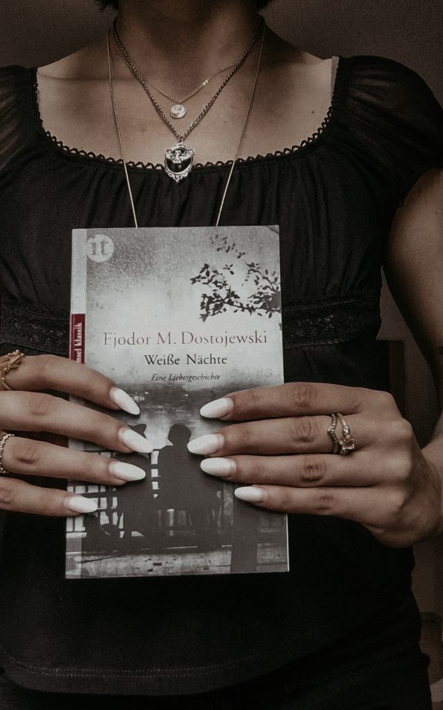 Vicky holding a book with a black an white cover with long white nails and gold rings on three fingers.