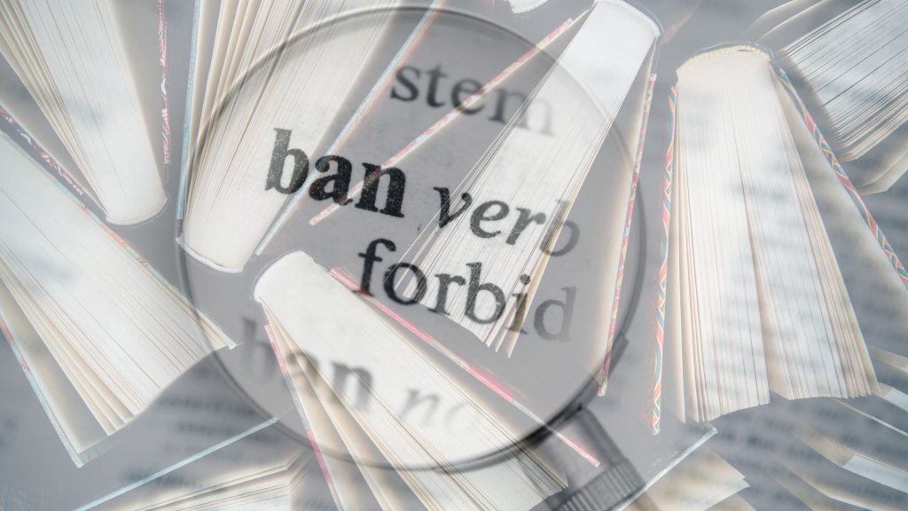 The dictionary definition of "ban" emphasized by a magnifying glass and a transparent image of books laid over it.