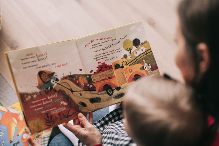 a woman reading to her child, child is sitting on her lap. a book is open and we can see a landscape and two tractors with eyes