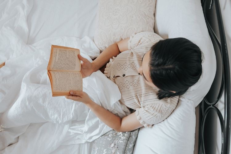 woman sitting in bed reading a book under the covers