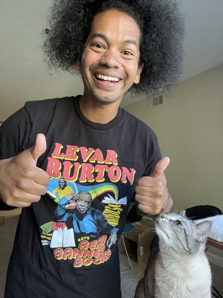 Mychal with thumbs up wearing a Levar Burton t-shirt that says "Levar Burton says read banned books" while a white and gray cat looks up at him
