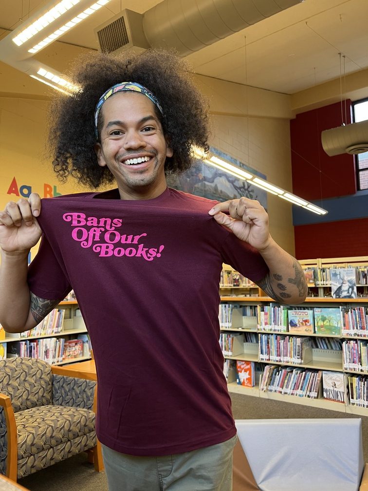 Mychal in a library while wearing a maroon t-shirt that states "Bans off our books!"