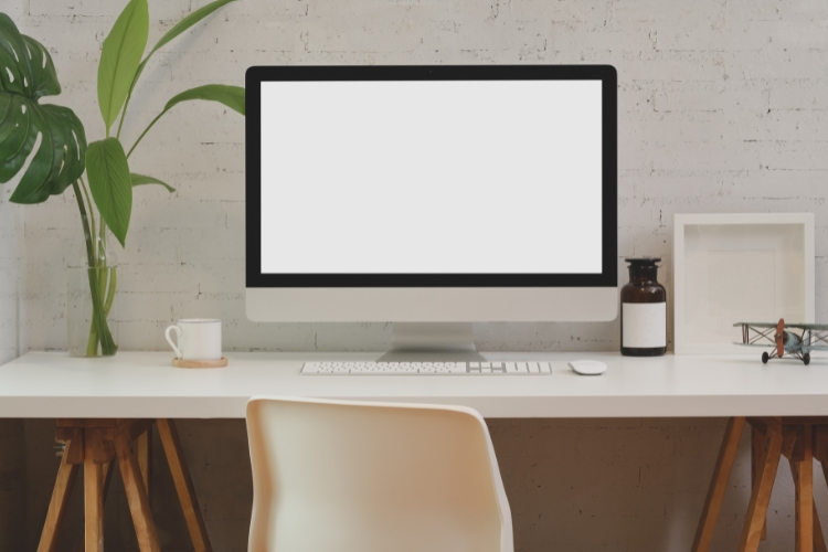 Blank computer screen on a white desk in front of a white brick wall.