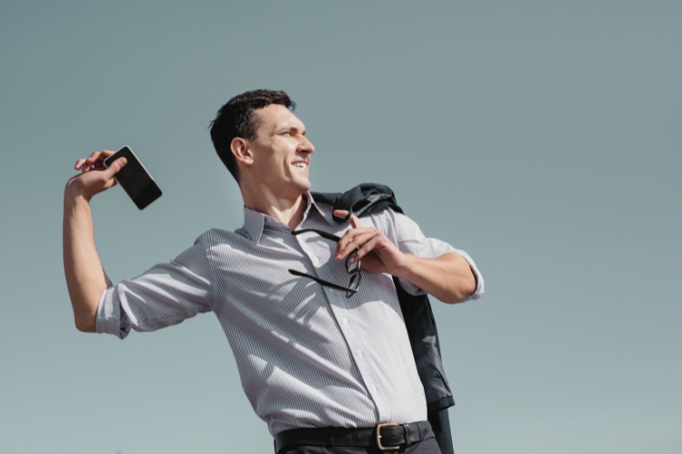 A man in a blue button-down shirt holds his glasses in one hand and his phone in the other, which is pulled back like he's going to throw it.